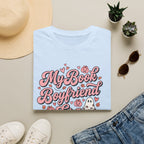 A folded light blue T-shirt with a pink and white graphic reading 'My Book Boyfriend' surrounded by hearts, pumpkins, and a ghost, styled in a flat lay with a straw hat, white sneakers, blue jeans, sunglasses, and small potted succulents on a beige background.