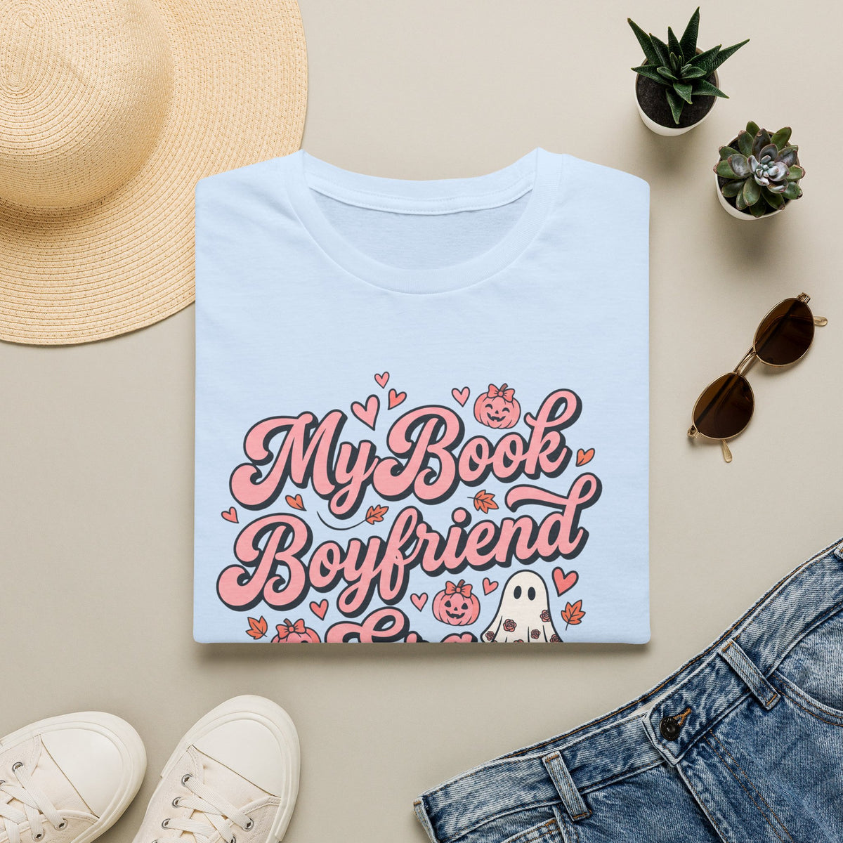 A folded light blue T-shirt with a pink and white graphic reading 'My Book Boyfriend' surrounded by hearts, pumpkins, and a ghost, styled in a flat lay with a straw hat, white sneakers, blue jeans, sunglasses, and small potted succulents on a beige background.