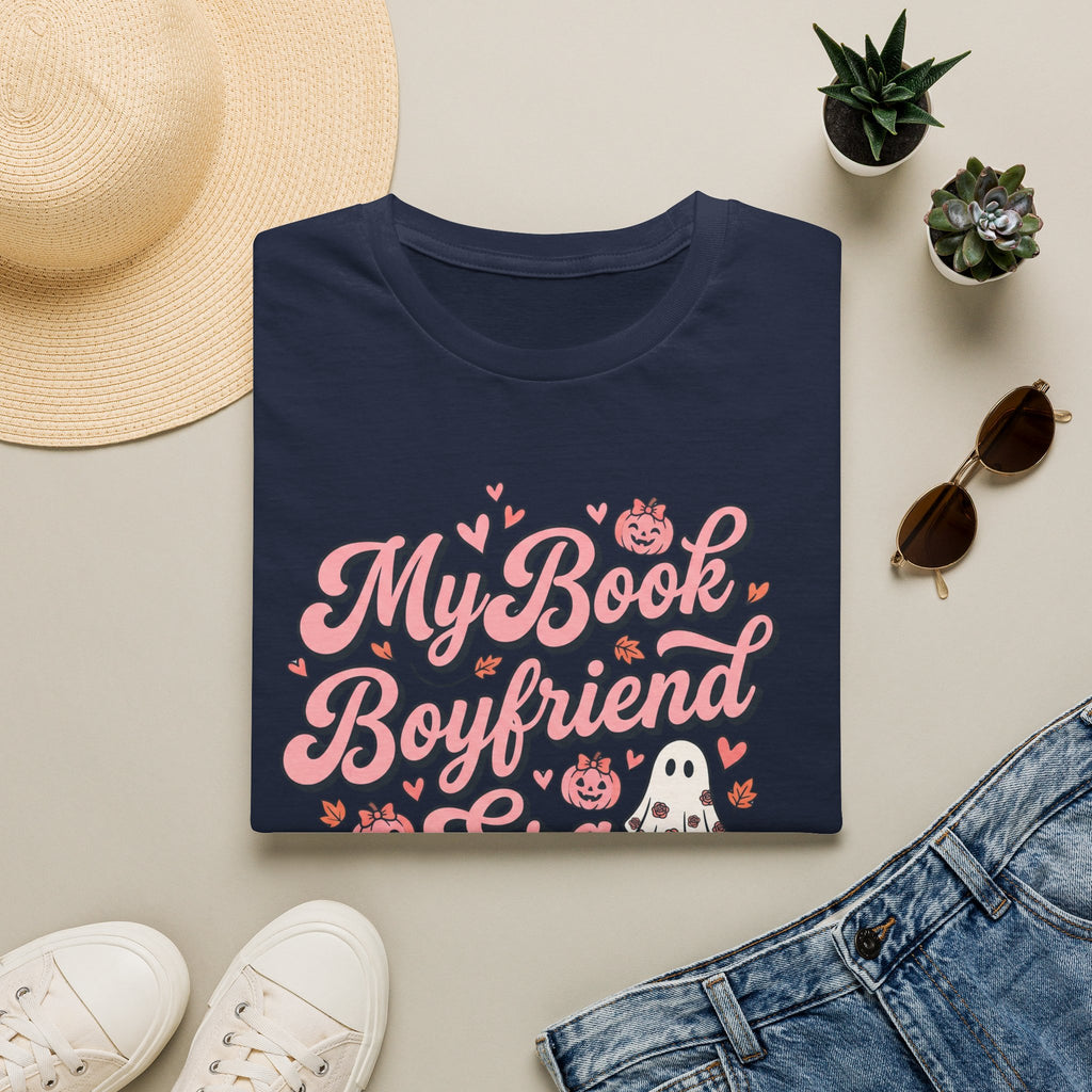 A navy blue folded T-shirt with a pink and white graphic reading 'My Book Boyfriend' surrounded by hearts, pumpkins, and a ghost, styled in a flat lay with a straw hat, small succulents, sunglasses, white sneakers, and blue jeans on a light beige background.