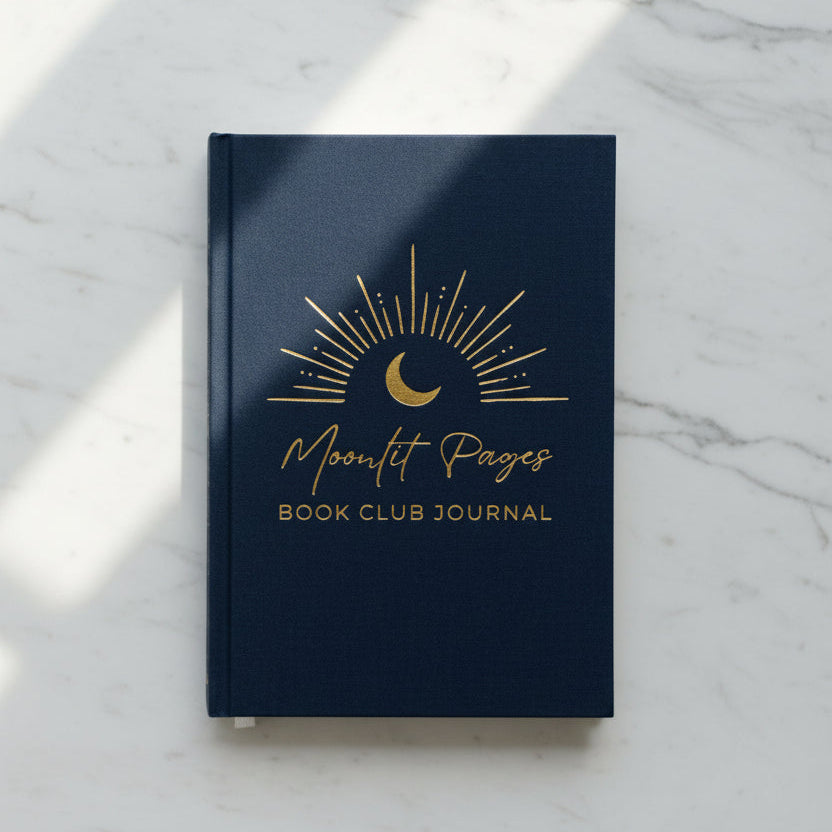 A navy blue hardcover book club journal with gold foil lettering and a crescent moon sunburst design, styled on a wooden tabletop next to a small potted plant and a white ceramic vase.