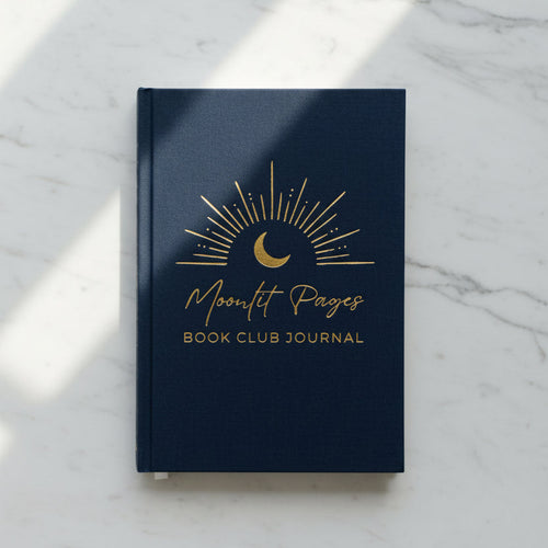 A navy blue hardcover book club journal with gold foil lettering and a crescent moon sunburst design, styled on a wooden tabletop next to a small potted plant and a white ceramic vase.