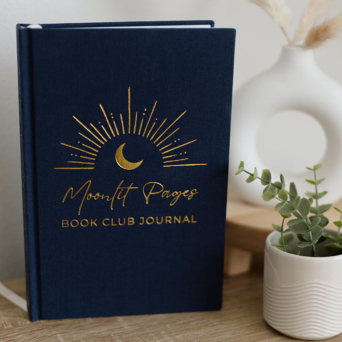 A navy blue hardcover book club journal with gold foil lettering and a crescent moon sunburst design, styled on a wooden tabletop next to a small potted plant and a white ceramic vase.