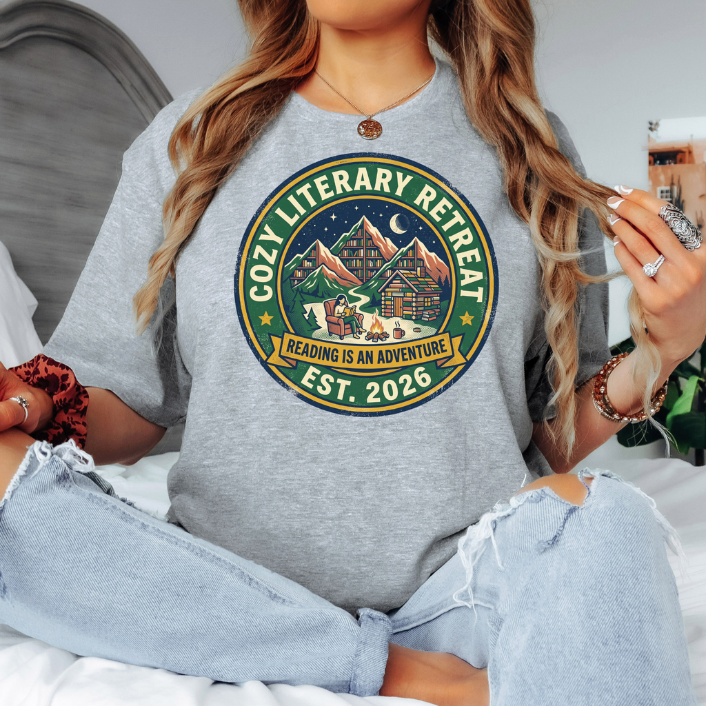 Heather gray t-shirt with a circular badge-style illustration of a person reading by a campfire outside a log cabin, with mountains shaped like bookshelves and a crescent moon in the night sky, text reading "Cozy Literary Retreat, Reading Is An Adventure, Est. 2026