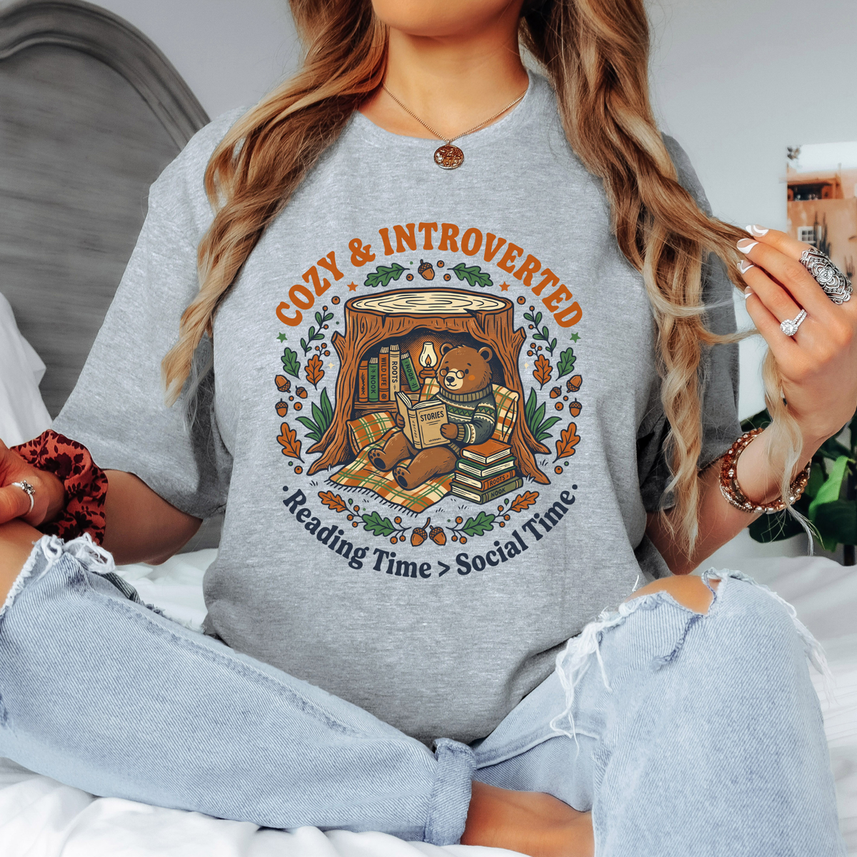Heather gray t-shirt with an illustration of a bear reading a book titled "Stories" inside a cozy tree stump nook, surrounded by autumn leaves and acorns, with text reading "Cozy & Introverted, Reading Time > Social Time.