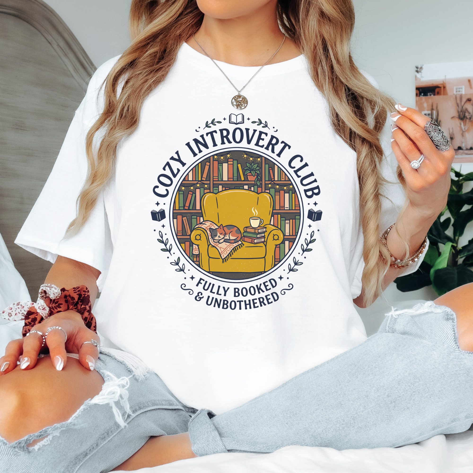 White t-shirt with a circular illustration of a sleeping calico cat curled up on a mustard yellow armchair next to a stack of books and a steaming mug, set against a cozy bookshelf background, with text reading "Cozy Introvert Club, Fully Booked & Unbothered.