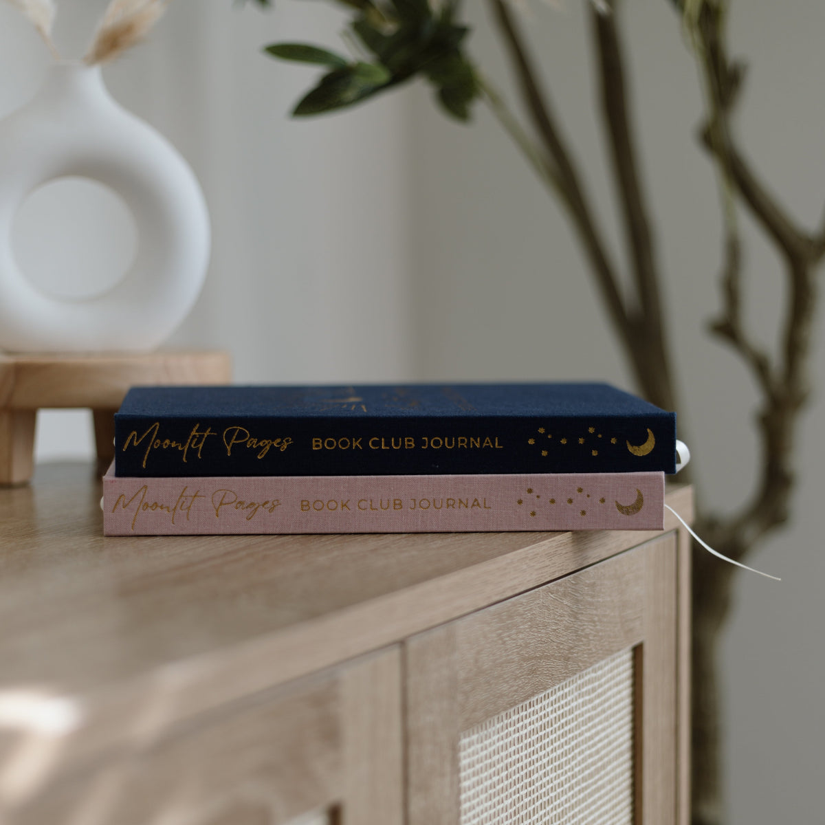Two hardcover journals stacked on a light wood cabinet in a styled interior setting. The top journal is dark navy blue with gold lettering, and the bottom journal is a dusty pink with gold lettering.