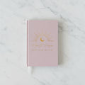A pink hardcover book club journal with gold foil text reading 'Moonlit Pages Book Club Journal' and a crescent moon sun design, styled on a wooden table with a small potted plant and a white vase in the background.