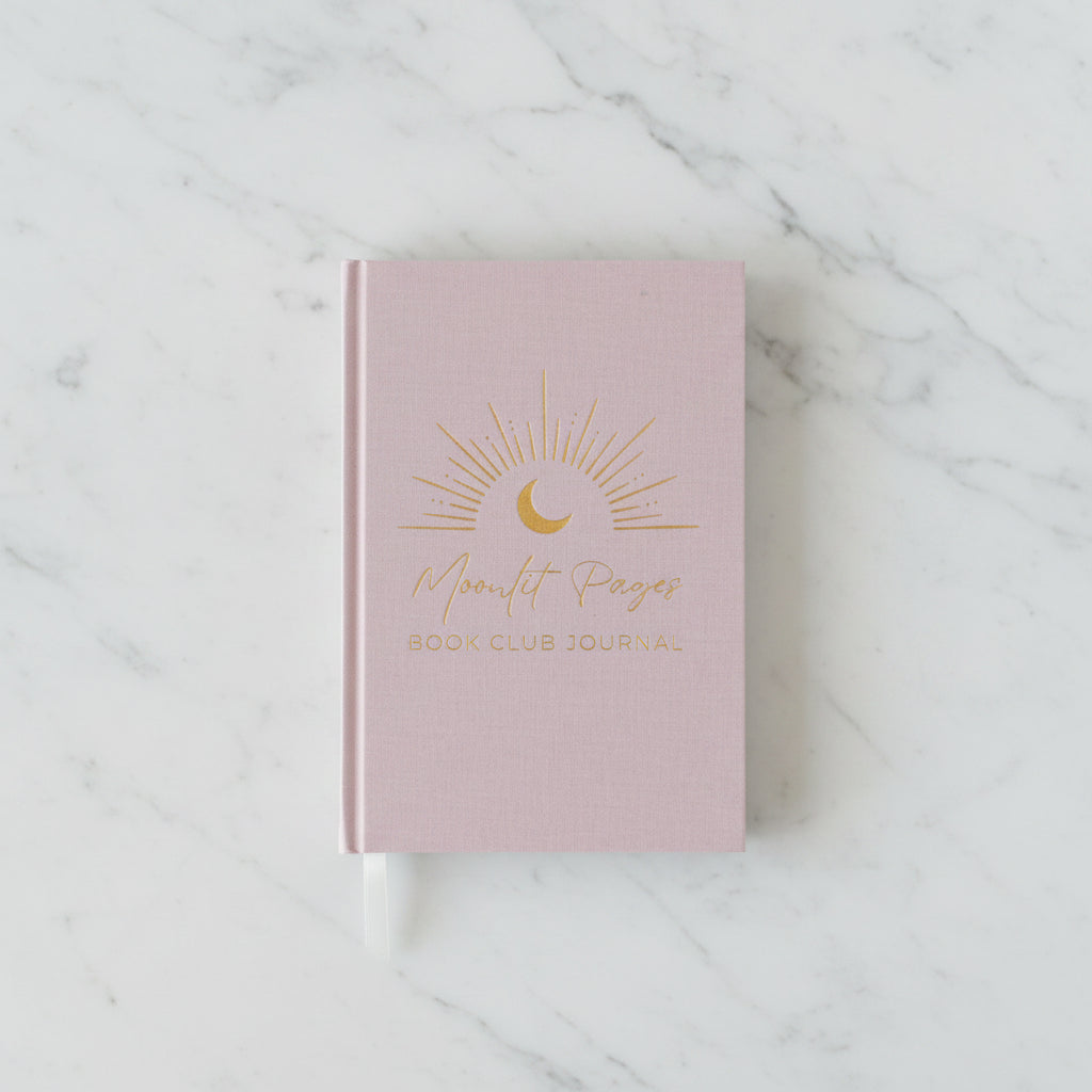 A pink hardcover book club journal with gold foil text reading 'Moonlit Pages Book Club Journal' and a crescent moon sun design, styled on a wooden table with a small potted plant and a white vase in the background.