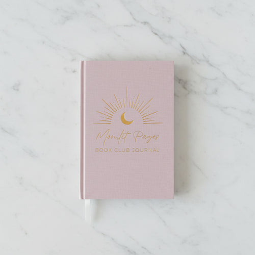 A pink hardcover book club journal with gold foil text reading 'Moonlit Pages Book Club Journal' and a crescent moon sun design, styled on a wooden table with a small potted plant and a white vase in the background.