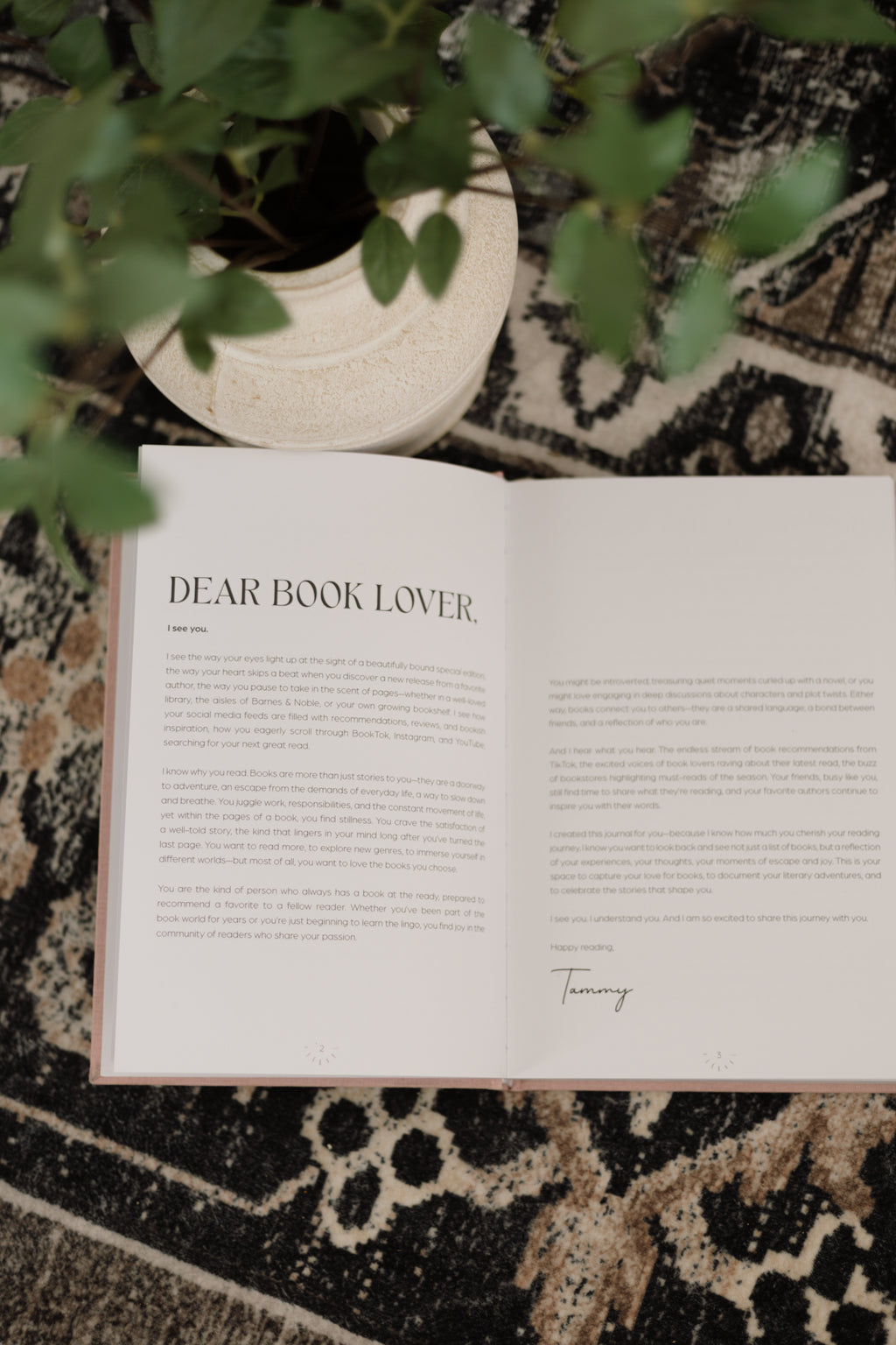 An open book with the title 'DEAR BOOK LOVER.' displayed on a patterned rug, styled with a potted plant in the background.