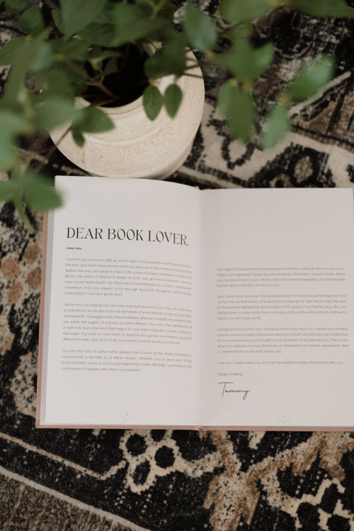 An open book with the title 'DEAR BOOK LOVER.' displayed on a patterned rug, styled with a potted plant in the background.