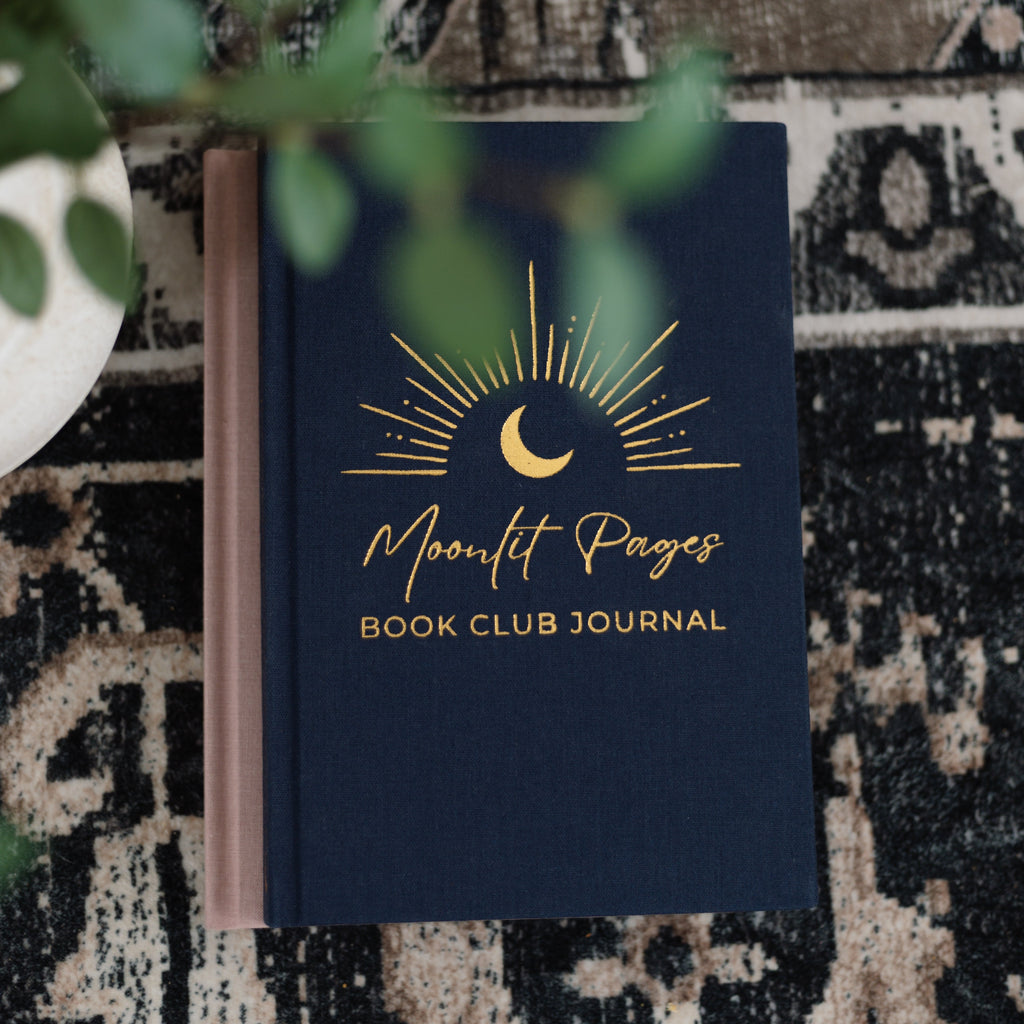 A navy blue book club journal with gold foil lettering and a crescent moon graphic, titled 'Moonlit Pages Book Club Journal'. The journal is styled on a black and beige patterned rug with green leaves in the foreground.