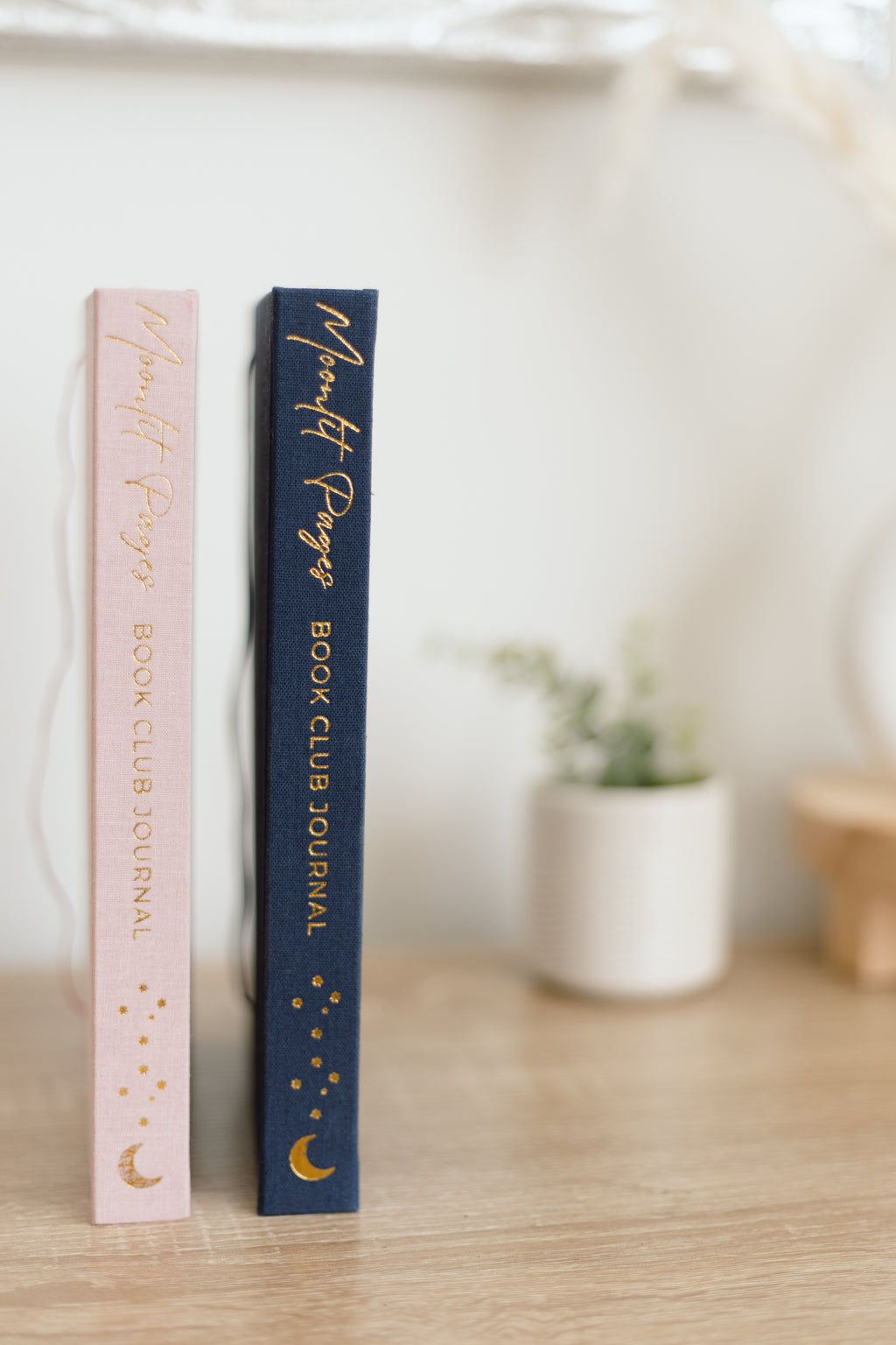 Two decorative book club journals standing upright on a wooden surface, one with a light pink cover and one with a dark navy cover, both featuring gold foil script and celestial designs, with a softly blurred indoor background.